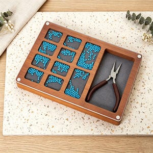May include: A wooden jewelry organizer with multiple compartments filled with turquoise beads. A section holds a pair of brown-handled pliers. The organizer sits on a speckled surface, suggesting a craft or jewelry-making theme.