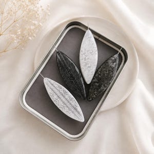 Puede incluir: Cuatro horquillas alargadas en forma de hoja dispuestas en una caja metálica rectangular. Dos horquillas son blancas con intrincados patrones geométricos y florales negros, y las otras dos son negras con detallados diseños texturizados en gris o blanco.