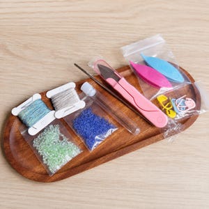 May include: A craft supply set featuring two spools of thread in blue and silver, two small bags of green and blue beads, a clear tube, pink thread snips, two plastic tatting shuttles in blue and pink, and several colorful plastic stitch markers.