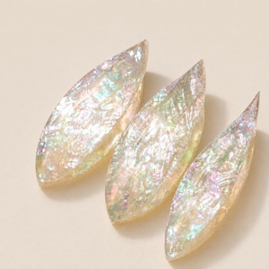 May include: Three marquise-shaped decorative pieces featuring a translucent, iridescent finish with embedded shimmering flakes that reflect light in shades of pink, green, and blue. Each piece has a smooth, curved surface and a pointed, tapered design.