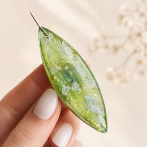 Op de afbeelding: Een marquise-vormige hanger van doorschijnende groene hars, met ingebedde witte bloemenelementen en kleine goudkleurige metaalschilfers door het materiaal heen. Een dunne zilverkleurige metalen pin steekt uit de bovenste punt.