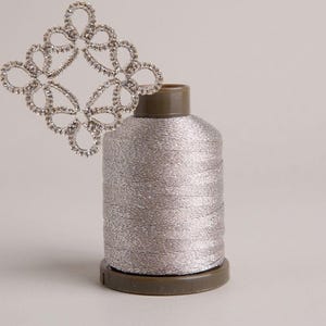 May include: A spool of metallic silver thread with a dark brown plastic core, positioned next to a decorative, diamond-shaped lace motif made of clear, sparkling rhinestones arranged in a repeating, intricate, looped pattern.