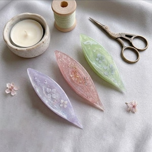 May include: Three shuttle tatting tools made of translucent resin in lavender, pink, and light green, each embedded with delicate dried flowers. The set includes a small white candle in a ceramic bowl, a wooden spool of thread, and metal scissors.