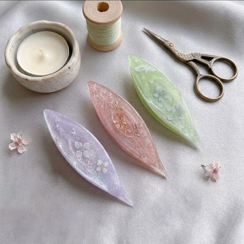 May include: Three shuttle tatting tools made of translucent resin in lavender, pink, and light green, each embedded with delicate dried flowers. The set includes a small white candle in a ceramic bowl, a wooden spool of thread, and metal scissors.