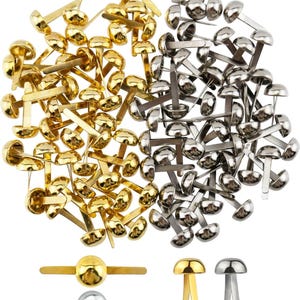 Op de afbeelding: Een verzameling goud- en zilverkleurige paperclips, ook wel brads genoemd, gerangschikt in een stapel. De brads hebben een afgeronde kop en twee platte, gespleten pinnen. Verschillende individuele brads worden eronder getoond.