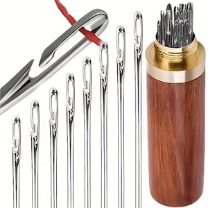 May include: A set of silver-coloured metal self-threading sewing needles with open-eye designs, displayed alongside a cylindrical wooden storage case with a brass-coloured metal screw-top lid, containing additional needles inside.