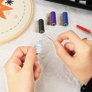May include: A silver-coloured metallic ornament being stitched with a needle and thread. Nearby are three small spools of thread in black, purple, and blue, along with an embroidery hoop featuring a star design and a red tool on a white fabric.