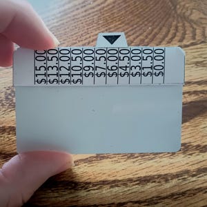 Puede incluir: Una etiqueta de precio blanca con una flecha negra y precios de 0,00 € a 15,00 €. La etiqueta está sujeta por una mano y está sobre una superficie de madera.