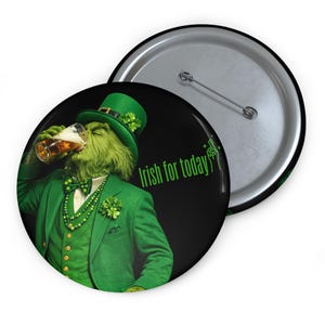 May include: A round button with a green Grinch character drinking beer, wearing a green suit and hat with shamrocks. The text "Irish for today" is printed in green on a black background. A silver pin back is visible.