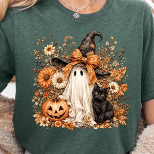 May include: A heather military green t-shirt featuring a graphic print of a white ghost wearing a black witch hat with an orange bow, a carved orange pumpkin, and a black cat surrounded by autumn-colored flowers and fall leaves.