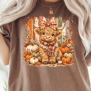 Op de afbeelding: Een bruin t-shirt met een afbeelding van een pluizige Schotse hooglander met een geruite strik en sjaal, omringd door herfstpompoenen, herfstbladeren en verticale penseelstreken in tinten oranje, groen en crème op een witte achtergrond.
