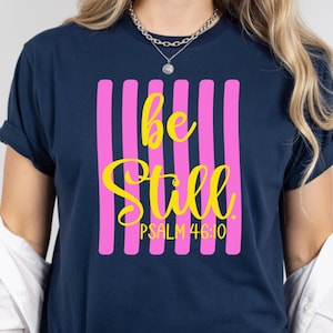 May include: Navy blue t-shirt with a vertical pink stripe design. The words "be Still" are in yellow script, with "PSALM 46:10" below. The shirt has rolled-up sleeves.