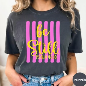 May include: Dark gray t-shirt with the words "be Still" in yellow script over vertical pink stripes. The text "PSALM 46:10" is below the main text. The shirt is a casual style.