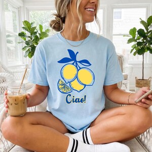 Op de afbeelding: Lichtblauw T-shirt met een citroenafbeelding en het woord "Ciao!" in het geel. De afbeelding toont drie gele citroenen en een schijfje citroen met blauwe bladeren. Het shirt is gemaakt van zacht materiaal.
