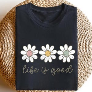 May include: Black t-shirt featuring three white daisy flowers with pink, yellow, and green centers. The phrase "life is good" is written in a cursive font below the flowers. The shirt is folded on a woven, light brown surface.