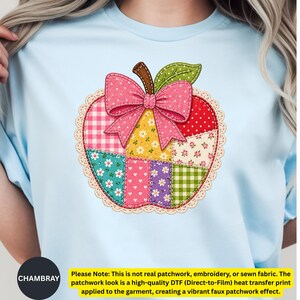 May include: A light blue t-shirt featuring a central graphic of an apple with a faux patchwork design. The apple is composed of various colourful patterned squares, including gingham, floral, and polka dot prints, topped with a pink polka dot bow.
