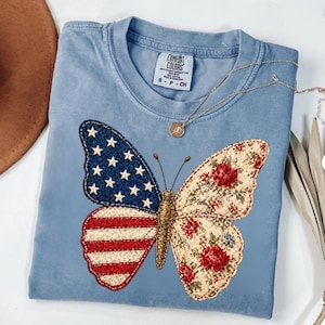Peut inclure: Un t-shirt bleu clair Comfort Colors avec un motif de papillon. Une aile affiche le drapeau américain, l'autre un motif floral avec des roses rouges. Un collier en or avec un pendentif est également visible.
