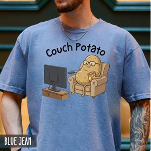Puede incluir: Una camiseta azul claro con las palabras "Couch Potato" sobre una patata de dibujos animados con gafas y sosteniendo un bocadillo, viendo la televisión. La camiseta también tiene las palabras "BLUE JEAN" en la parte inferior.