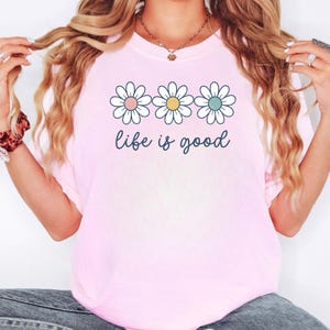May include: A light pink t-shirt featuring three daisy flowers with pink, yellow, and blue centers. The words "life is good" are written in a cursive font below the flowers. The shirt is a casual style.