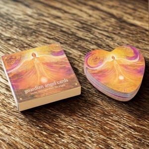 Pode incluir: Um baralho de "cartas de anjos da guarda" e uma pilha de cartas em forma de coração. As cartas apresentam uma ilustração de um anjo em tons de laranja, amarelo e roxo. O texto na caixa diz "cartas de anjos da guarda mensagens de amor dos anjos."