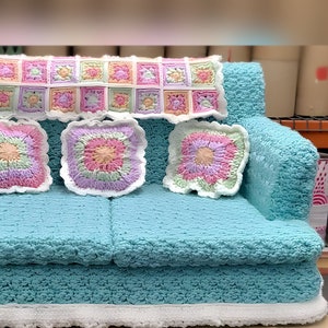 Könnte beinhalten: Ein türkisfarbenes Häkelsofa mit drei quadratischen Kissen und einem rechteckigen Überwurf. Die Kissen und der Überwurf weisen pastellfarbene Blumenmuster mit weißen Rändern auf. Das Sofa hat einen weißen Häkelbesatz am Sockel.