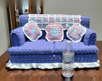 Könnte beinhalten: Ein violettes, gehäkeltes Miniatursofa mit weißem Rüschensaum, drei quadratischen Granny-Square-Kissen in Rosa, Blau und Weiß sowie einer passenden Decke über der Rückenlehne, platziert neben einer durchsichtigen Plastikwasserflasche.