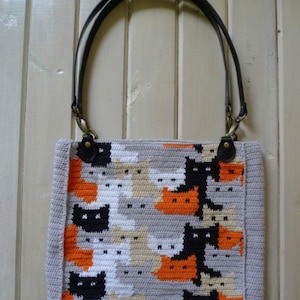 May include: A crocheted tote bag with a pattern of black, white, orange, and gray cats. The bag has black leather straps with brass hardware.