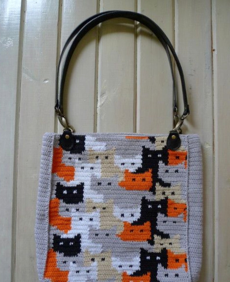 May include: A crocheted tote bag with a pattern of black, white, orange, and gray cats. The bag has black leather straps with brass hardware.