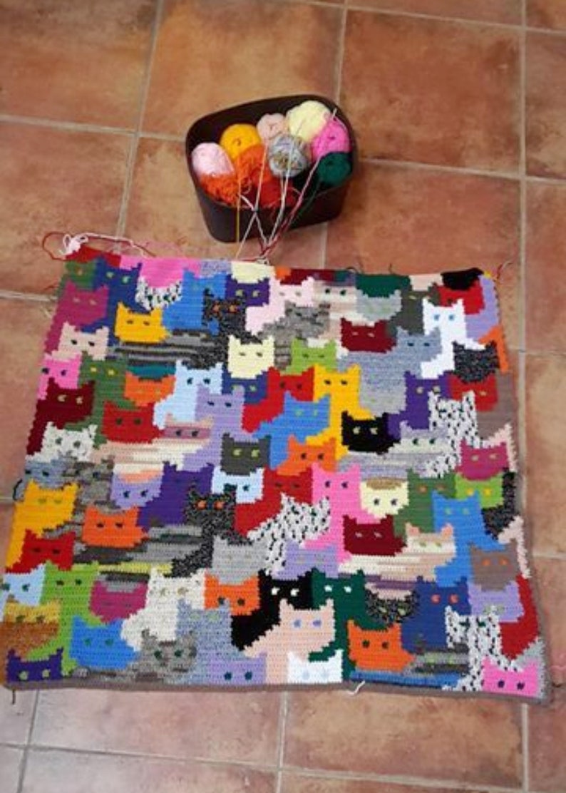 May include: A colorful crocheted blanket featuring a pattern of cats in various colors and designs. The blanket is made with different colored yarn and is a unique and handmade piece.