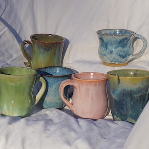 Può includere: Una collezione di sei tazze in ceramica fatte a mano in vari colori. Le tazze presentano forme e texture uniche, con colori tra cui verde, blu, rosa e tonalità della terra. Ogni tazza ha un manico e un aspetto rustico e artigianale.