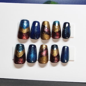 Pode incluir: Dez unhas artificiais dispostas em duas fileiras. Cada unha tem uma cor e design diferentes, incluindo azul, dourado e bordô. Algumas unhas têm um design de chevron. As unhas estão em suportes brancos.