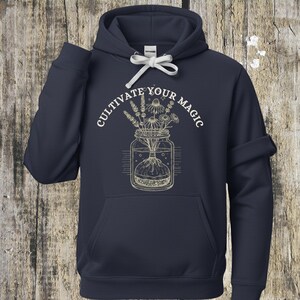 Puede incluir: Sudadera con capucha azul marino con cordón blanco y bolsillo delantero. La sudadera presenta un gráfico de un tarro lleno de flores y el texto "Cultivate Your Magic" en fuente curva.