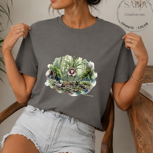Puede incluir: Una camiseta gris con una ilustración de acuarela de una orquídea cara de mono en un entorno forestal. La camiseta tiene mangas cortas y cuello redondo. El texto "SABIA Creative lab. GRANITE : COLOR" está en la esquina superior derecha.