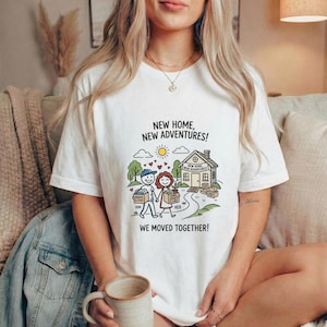 以下が含まれることがあります： 新しい家に引っ越すカップルのグラフィックが描かれた白いTシャツ。デザインには家、木、太陽、そして「NEW HOME, NEW ADVENTURES!」と「WE MOVED TOGETHER!」というテキストが含まれています。