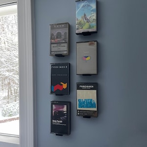 Può includere: Una collezione di sette musicassette montate su una parete azzurra. I nastri presentano copertine di album di band come Rush, Foreigner e Deep Purple. Ogni nastro è esposto in un supporto nero, creando un display musicale retrò.