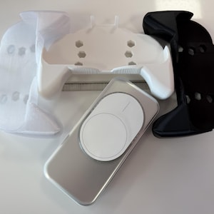 Pode incluir: Uma variedade de acessórios impressos em 3D em branco, preto e prata. Os itens incluem um dispositivo semelhante a um telefone com um design circular branco e vários componentes em forma de controlador. Os objetos estão dispostos em uma superfície branca.