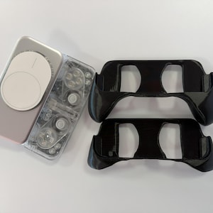 May include: A rectangular electronic device with a clear casing and a white circular magnetic attachment, positioned next to two black, contoured plastic controller grips featuring cutouts for buttons and joysticks on the front surface.