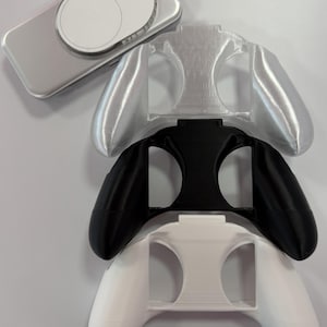 Puede incluir: Un surtido de accesorios para mandos de videojuegos en plata, negro y blanco. La imagen muestra una funda plateada con un panel de control circular y tres empuñaduras de mando de diferentes colores, apiladas.