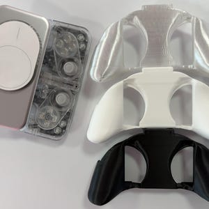 May include: A handheld gaming device with a transparent casing and a white circular magnetic attachment, accompanied by three separate controller grip attachments in clear, solid white, and solid black colors, arranged vertically to the right.