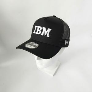 May include: Black baseball cap with the white letters "IBM" on the front. The cap has a curved brim and a mesh back. The New Era logo is on the side and the "9FORTY" sticker is on the brim.