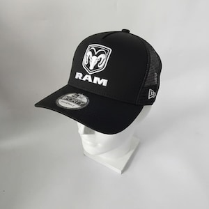 May include: Black trucker hat with a white Ram logo and the word "RAM" embroidered on the front. The hat has a curved brim and a mesh back. A "9FORTY" sticker is on the brim. The hat is displayed on a white mannequin head.