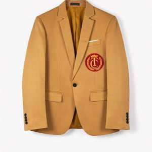 Puede incluir: Una blazer color beige con un emblema rojo y dorado en el pecho izquierdo. La blazer tiene un cierre de botón negro, dos bolsillos delanteros y cuatro botones negros en cada manga. El forro interior es de color amarillo dorado.