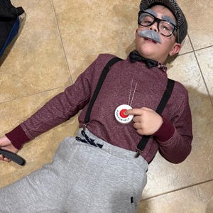 May include: A person dressed in an old man costume, wearing a burgundy sweater, gray sweatpants, suspenders, a bow tie, glasses, a gray mustache, and a newsboy cap. A button that says "PUSH FOR HELP" hangs from a string.