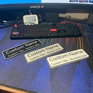 May include: A black mechanical keyboard with red and purple keycaps, a small white device with colorful buttons, and three name tags with the text "Custom Name" and "Custom Title" in different fonts. The tags are black and white.