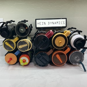 May include: A display of various water bottles and tumblers in a black rack. The bottles come in a variety of colors, including black, yellow, orange, and white. The text "HEIN DYNAMICS" is displayed on a small illuminated sign.