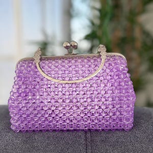 May include: A lavender beaded handbag with a silver clasp and handle. The rectangular purse is covered in small, clear beads, giving it a textured appearance. The handle is a curved, silver metal. The clasp is a silver metal ball.