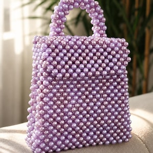 Könnte beinhalten: Eine strukturierte Handtasche, die vollständig mit kleinen, glänzenden lavendelfarbenen Perlen bedeckt ist. Die Tasche hat einen rechteckigen Körper mit Klappverschluss und einen einzelnen abgerundeten Tragegriff aus einheitlichen, violetten Perlen.