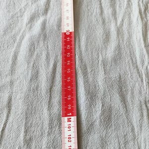 K&ouml;nnte beinhalten: Ein rot-wei&szlig;es Ma&szlig;band, in Zentimetern markiert, liegt vertikal auf einem hellgrauen Stoff. Das Band zeigt Messungen von 82 cm bis 104 cm, mit deutlichen Markierungen und Zahlen.
