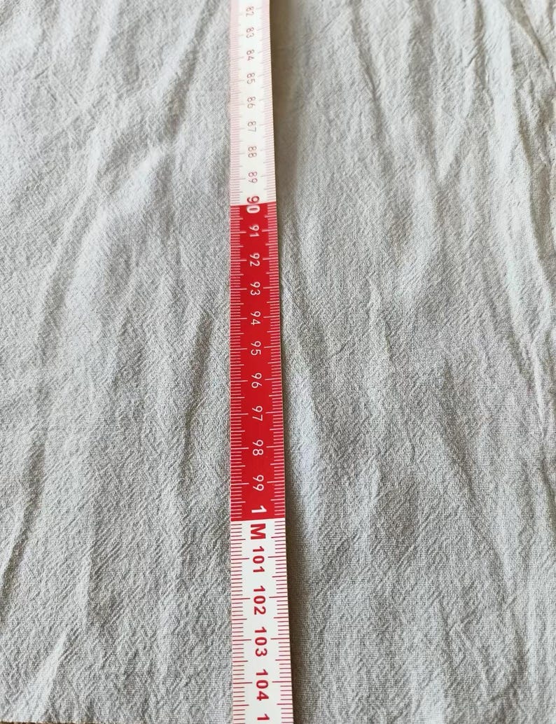 K&ouml;nnte beinhalten: Ein rot-wei&szlig;es Ma&szlig;band, in Zentimetern markiert, liegt vertikal auf einem hellgrauen Stoff. Das Band zeigt Messungen von 82 cm bis 104 cm, mit deutlichen Markierungen und Zahlen.