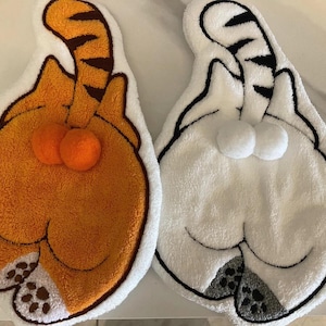 May include: Two cat-shaped rugs. One is orange with a brown striped tail and two orange pom-poms. The other is white with a black striped tail and two white pom-poms. Both have paw prints.